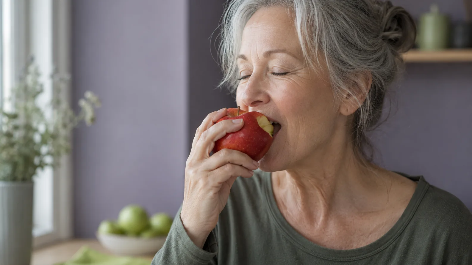 Woman with grey hair biting into a red apple with her eyes closed