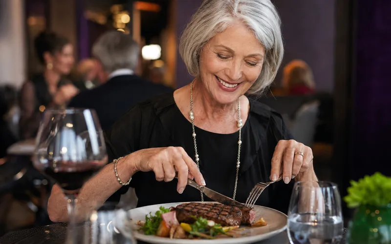 Woman in her sixties smiling while cutting a steak at a restaurant table